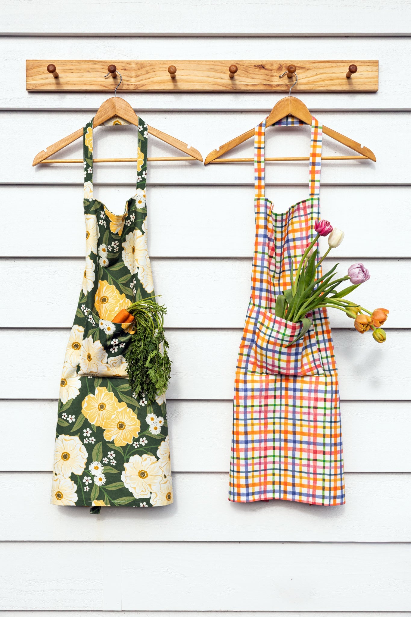 Two colorful aprons hanging on wooden hangers from a wooden peg rack against a white wood-plank wall. The left apron has a green, yellow, and white floral print with a pocket holding carrots and greens.