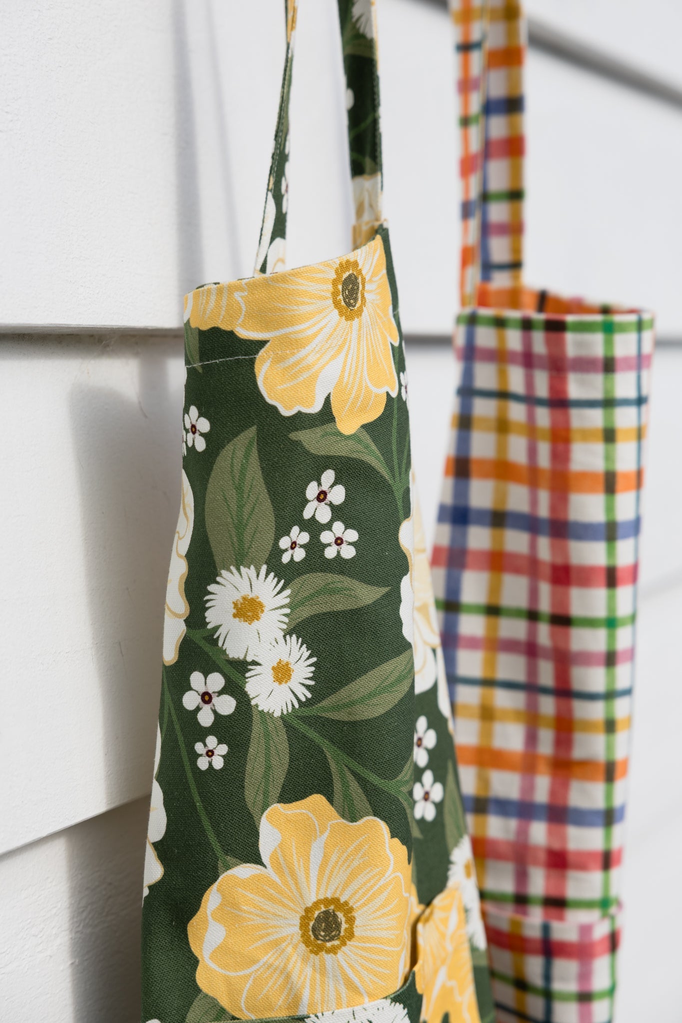 A dark green apron with a yellow and white floral print hanging on a white paneled wall, with a second multicolor plaid apron partially visible behind it.