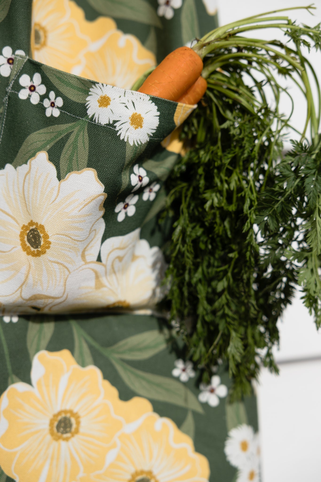 A close-up of a dark green apron with a large floral print of white daisies and yellow flowers, featuring a side pocket holding a fresh carrot with green tops.