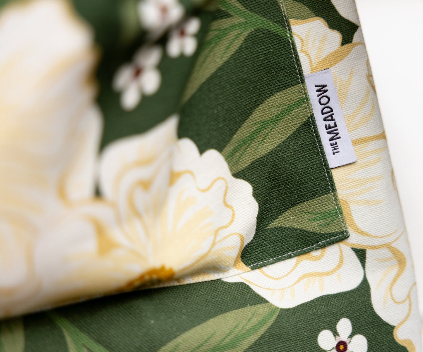 Close-up of a fabric product with a green, white, and yellow floral pattern, featuring a white fabric tag with the brand name 'THE MEADOW'.